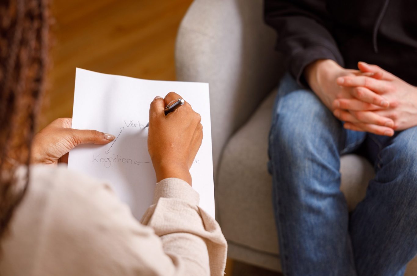 Therapiesitzung Eine Therapeutin schreib "Kognition, Verhalten" auf einen Zettel. Ein:e Patient:in sitzt mich gefalteten Händen gegenüber